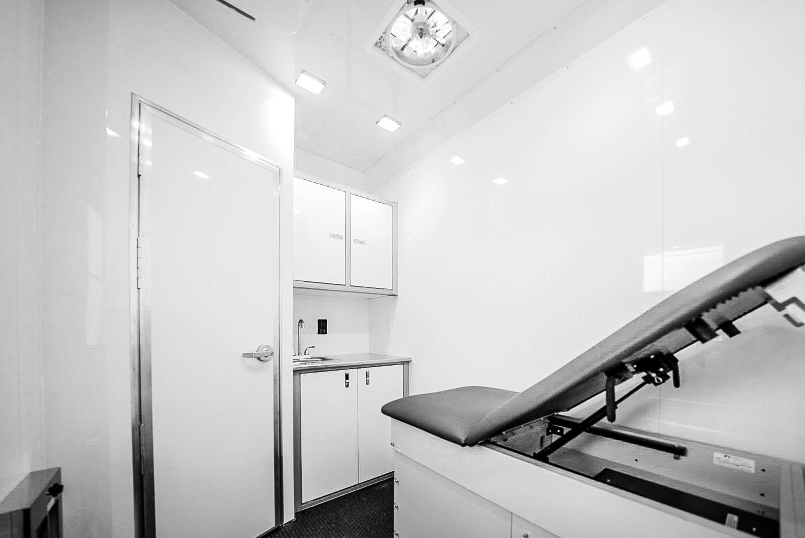 Interior of a Wellness on Wheels mobile healthcare unit, featuring a clean, stark white exam room with a door, sink, and exam table.